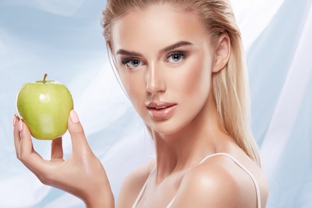 Beautiful girl with nude make up and blonde hair posing with green apple at bright backgroundの写真素材