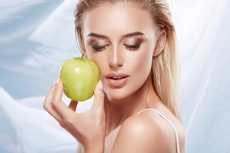 Beautiful girl with nude make up and blonde hair posing with green apple at bright backgroundの写真素材