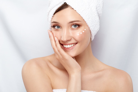 young and beautiful female with towel on her head and cream on the cheek smiling and touching her face with one arm, face treatment conceptの写真素材