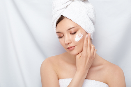 beautiful woman with perfect clean skin applying cream on her face after bath, pretty girl in bath towel moisturizing cheeksの写真素材
