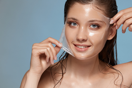 woman peels-off transparent facial mask and smiling to the camera on blue, happy anf fresh girl with perfect skinの写真素材