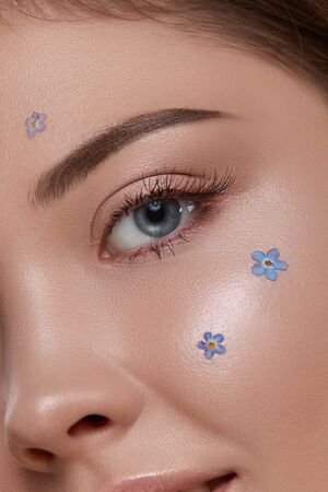 romantic woman's eye looking to the camera with fresh blue flowers on cheek, close-up of woman face with forget-me-nots and shiny muaの写真素材