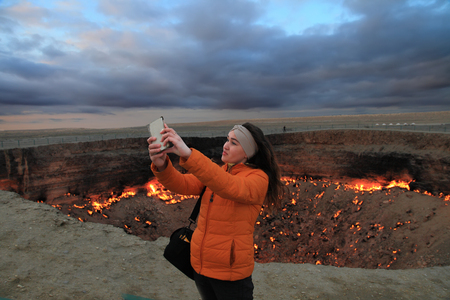 Turkmenistan, Darvaza - March 21, 2019:   Girl makes selfie on the background of the burning crater "Gates of Hell", Karakum desert. Darvaza is one of the ten most unusual places in the world.のeditorial素材