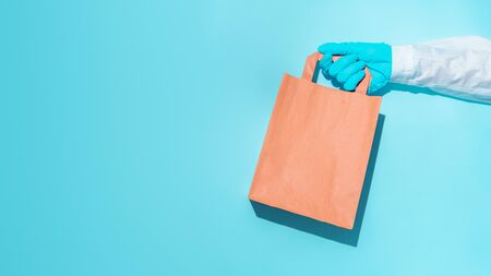 hand in gloves holding shopping paper bag. Concept of consumerism, shopping symbol during pandemic time. Copy space trend pastel color. Hard shadows, morning light.の写真素材