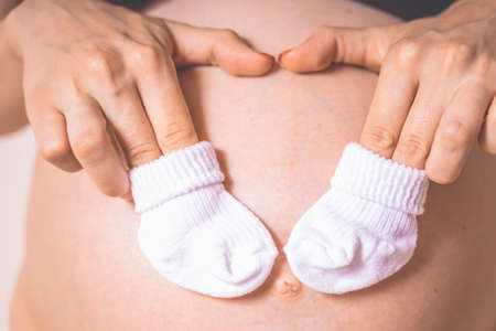 Pregnant woman with clothing for newborn baby close up. White woolen little baby slippers, socks in...