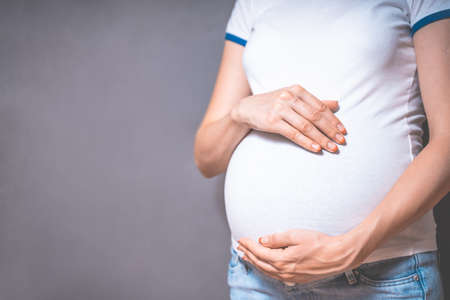 Pregnant woman holds hands on belly on a gray background. Copy space, pregnancy, motherhood, people and expectation concept. Close-up, copy space, indoors. Toned photo of pregnancy.の写真素材