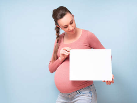 Young beautiful pregnant woman over isolated blue background holding a blank sheet of paper....