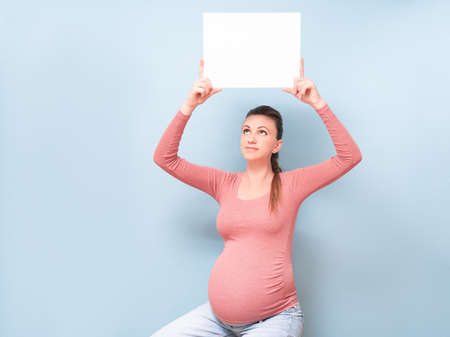 Young beautiful pregnant woman over isolated blue background holding a blank sheet of paper. Lifestyle Concept, your text here. Copy spaceの写真素材
