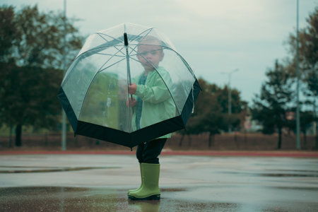 Children playing outside summer rain time with umbrella. Boy is wearing green raincoat and Rubber boots. Kid having fun at rainy day. Childchood, autumn conceptの写真素材
