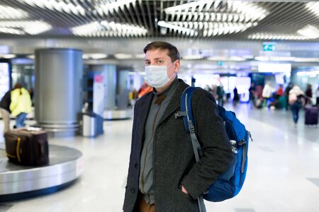Young European man in gray coat, protective disposable medical mask in airport. Afraid of dangerous N-CoV 2019 influenza coronavirus mutated and spreading in China. Blue backpack, suitcase on wheels.の写真素材