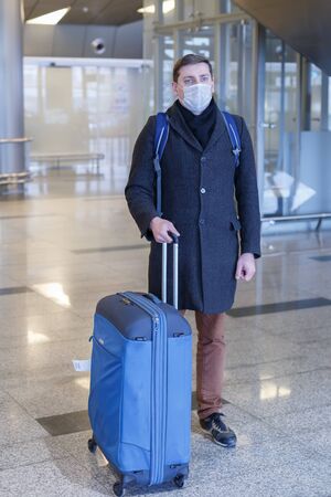 Young European man in gray coat, protective disposable medical mask in airport. Afraid of dangerous N-CoV 2019 influenza coronavirus mutated and spreading in China. Blue backpack, suitcase on wheels.の写真素材