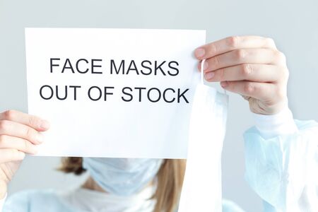 Woman in disposable blue medical shirt holds in hands information plate with text: face masks are out of stock, sold out. Epidemic of N-CoV 2019 influenza coronavirus mutated and spreadingの写真素材