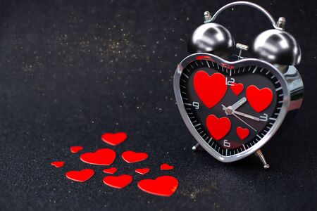 Steel Clock alarm in the form of a heart with red hearts inside and on the table on a black shiny background. Happy Valentine's Dayの写真素材