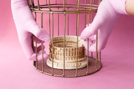 Stone statuette of the Coliseum building in the city of Rome in Italy, enclosed in a golden cage. Hands of doctor in medical gloves. Dangerous infection of coronavirus covid-2019. Pink background.の写真素材