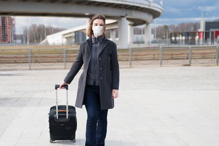 Stylish scared woman in protective face mask, gray wool coat and suitcase on the street near metro station leaves the city. Self protection in transport during quarantine from coronavirus covid-2019.の写真素材