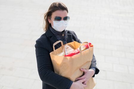 Scared woman in protective face mask,gray coat.Making store of food in craft bag before isolation:toilet paper,paste,cookies.Lack of products.Leaving city during quarantine of coronavirus covid-2019.の写真素材