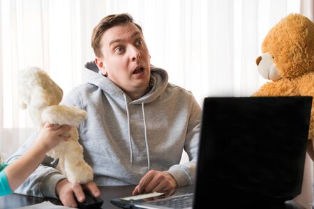 Young man in sports hoody at telework due to home quarantine of coronovirus pandemic COVID-2019.Children interfere remote work.Soft toys: bear, sheep.Online conferencen on headset with boss by laptop.の写真素材