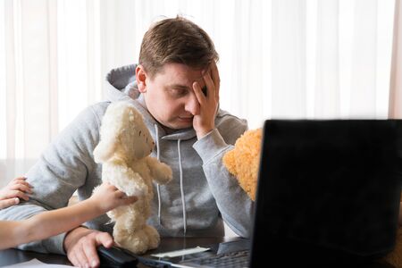 Young man in sports hoody at telework due to home quarantine of coronovirus pandemic COVID-2019.Children interfere remote work.Soft toys: bear, sheep.Online conferencen on headset with boss by laptop.の写真素材