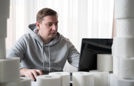 Young man in sports hoody switched to telework due to quarantine of coronovirus pandemic COVID-2019. Remote work at home table with toilet paper. Communication with boss by laptop. Online conference.の写真素材