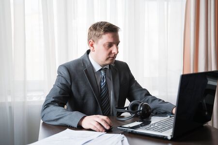 Young man in gray suit, shirt at telework due to quarantine of coronovirus pandemic COVID-2019.Remote work at home table.Communication on headset with boss,colleagues through laptop.Online conference.の写真素材