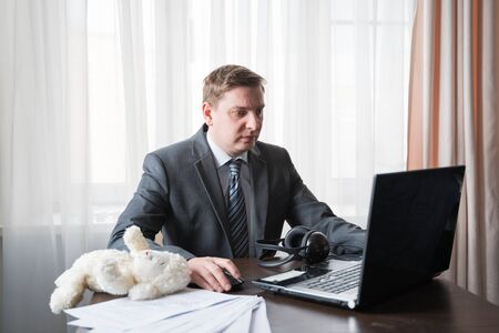 Young man in gray suit, shirt at telework due to quarantine of coronovirus pandemic COVID-2019. Remote work at home table with children's soft toy. Online conferencen on headset with boss by laptop.の写真素材