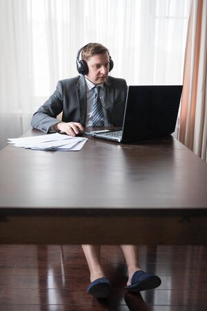 Young man in gray suit, shirt with bare feet in slippers at telework due to quarantine of coronovirus pandemic COVID-2019.Remote work at home table.Online conference on headset by laptop with boss.の写真素材
