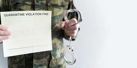 Police officer with handcuffs in khaki colored uniform stops people on the street for violating rules of quarantine of home self-isolation due to coronovirus covid-19 flu pandemic.Yellow fine receipt.の写真素材