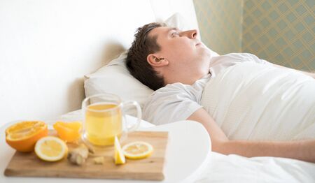 Cup with antipyretic drugs for colds,flu.Sick man in bed. Tea with citrus vitamin C,ginger root,lemon,orange.Wooden tray. Home self-treatment.Medical quarantine antiviral covid-19 coronavirus therapy.の写真素材