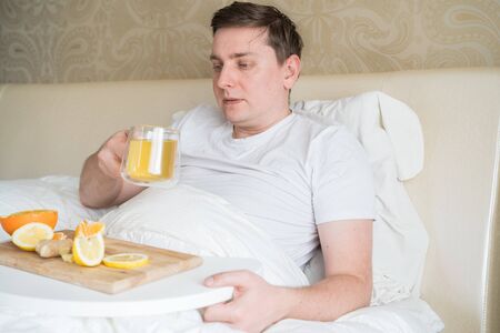 Cup with antipyretic drugs for colds,flu.Sick man in bed. Tea with citrus vitamin C,ginger root,lemon,orange.Wooden tray. Home self-treatment.Medical quarantine antiviral covid-19 coronavirus therapy.の写真素材