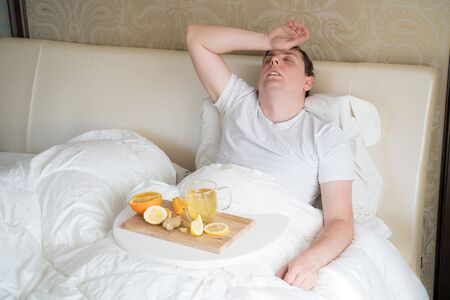 Cup with antipyretic drugs for colds,flu.Sick man in bed. Tea with citrus vitamin C,ginger root,lemon,orange.Wooden tray. Home self-treatment.Medical quarantine antiviral covid-19 coronavirus therapy.の写真素材