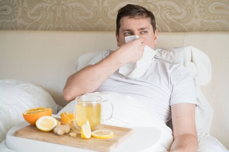 Cup with antipyretic drugs for colds,flu.Sick man in bed. Tea with citrus vitamin C,ginger root,lemon,orange.Wooden tray. Home self-treatment.Medical quarantine antiviral covid-19 coronavirus therapy.の写真素材