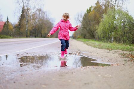 Happy little girl in pink waterproof jacket, rubber boots cheerfully jumps through puddles on street road in rainy weather. Spring, autumn. Children's fun in fresh air after rain. Outdoors recreation.の写真素材