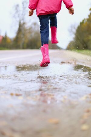 Happy little girl in pink waterproof jacket, rubber boots cheerfully jumps through puddles on street road in rainy weather. Spring, autumn. Children's fun in fresh air after rain. Outdoors recreation.の写真素材