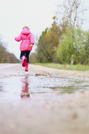 Happy little girl in pink waterproof jacket, rubber boots cheerfully jumps through puddles on street road in rainy weather. Spring, autumn. Children's fun in fresh air after rain. Outdoors recreation.の写真素材