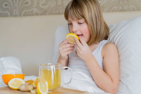 Sick girl in bed tastes lemon slice.Cup of antipyretic drugs for colds,flu.Tea with citrus vitamin C,ginger root,orange.Wooden tray.Home self-treatment.Medical quarantine covid-19 coronavirus therapy.の写真素材