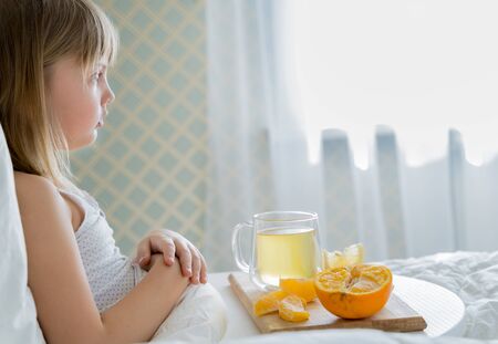 Little sick girl in bed with cup of antipyretic drugs for colds,flu.Tea with citrus vitamin C,ginger root,lemon,orange.Wooden tray. Home self-treatment.Medical quarantine covid-19 coronavirus therapy.の写真素材