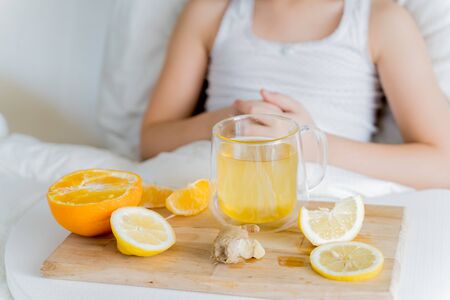 Little sick girl in bed with cup of antipyretic drugs for colds,flu.Tea with citrus vitamin C,ginger root,lemon,orange.Wooden tray. Home self-treatment.Medical quarantine covid-19 coronavirus therapy.の写真素材