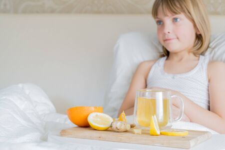 Little sick girl in bed with cup of antipyretic drugs for colds,flu.Tea with citrus vitamin C,ginger root,lemon,orange.Wooden tray. Home self-treatment.Medical quarantine covid-19 coronavirus therapy.の写真素材