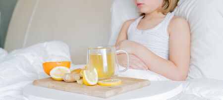 Little sick girl in bed with cup of antipyretic drugs for colds,flu.Tea with citrus vitamin C,ginger root,lemon,orange.Wooden tray. Home self-treatment.Medical quarantine covid-19 coronavirus therapy.の写真素材