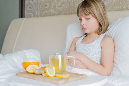Little sick girl in bed with cup of antipyretic drugs for colds,flu.Tea with citrus vitamin C,ginger root,lemon,orange.Wooden tray. Home self-treatment.Medical quarantine covid-19 coronavirus therapy.の写真素材