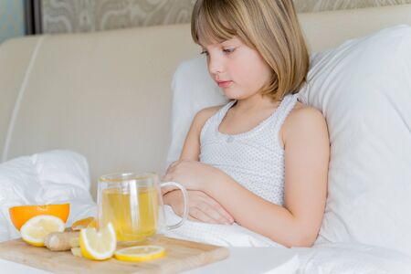 Little sick girl in bed with cup of antipyretic drugs for colds,flu.Tea with citrus vitamin C,ginger root,lemon,orange.Wooden tray. Home self-treatment.Medical quarantine covid-19 coronavirus therapy.の写真素材