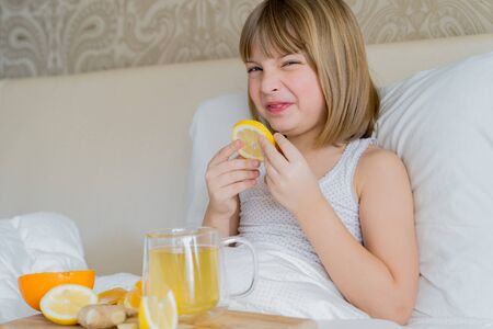 Sick girl in bed tastes lemon slice.Cup of antipyretic drugs for colds,flu.Tea with citrus vitamin C,ginger root,orange.Wooden tray.Home self-treatment.Medical quarantine covid-19 coronavirus therapy.の写真素材