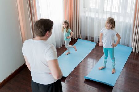 Home workout in flat.Dad teaching little girls fitness, daughters. Physical morning exercises on blue yoga mats.Family children fun.Healthy indoor activity.Quarantine,coronavirus covid-19.No equipment.の写真素材