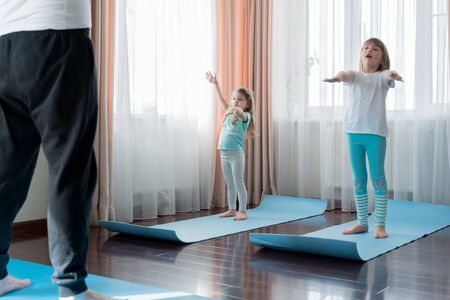 Home workout in flat.Dad teaching little girls fitness, daughters. Physical morning exercises on blue yoga mats.Family children fun.Healthy indoor activity.Quarantine,coronavirus covid-19.No equipment.の写真素材