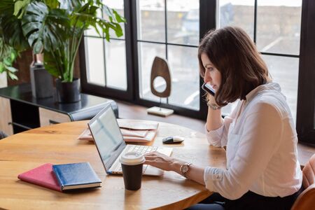 Business woman in white shirt working at laptop at home office near big window,drinking...