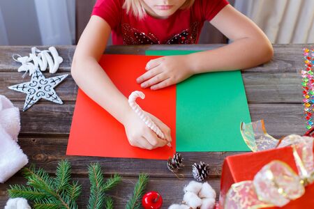 Writing letter to Santa Claus on red, green paper with candy cane shaped pink pen. Little girl writes wishes, Christmas gifts.Hat,toys,cones,fur tree branch,books,cotton flowers, presents,snow apples.の写真素材