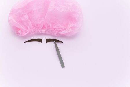 Disposable medical hat for beauty salon customers and patients. Decorative carved brown brows, tweezers on a pink background.Flat lay: accessories for coloring, plucking and shaping eyebrows.の写真素材
