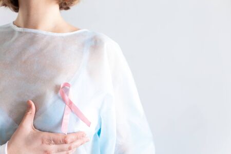 International cancer day. A woman with short blond hair, in a disposable medical blue shirt, in a white bra holds her hand over her breast. Pink ribbon.の写真素材
