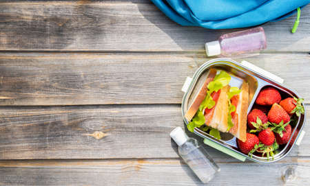 Lunchbox with sandwiches, fresh strawberries, two bottles of sanitizer and blue backpack on wooden background. Back to school after quarantine. Lunch break, box with safety precautions after coronavirus.の写真素材