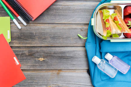 Lunchbox with sandwiches. Box of fresh strawberries, bottles of sanitizer and blue backpack with notebooks and pens on wooden background.Back to school.Lunch with safety precautions after coronavirusの写真素材
